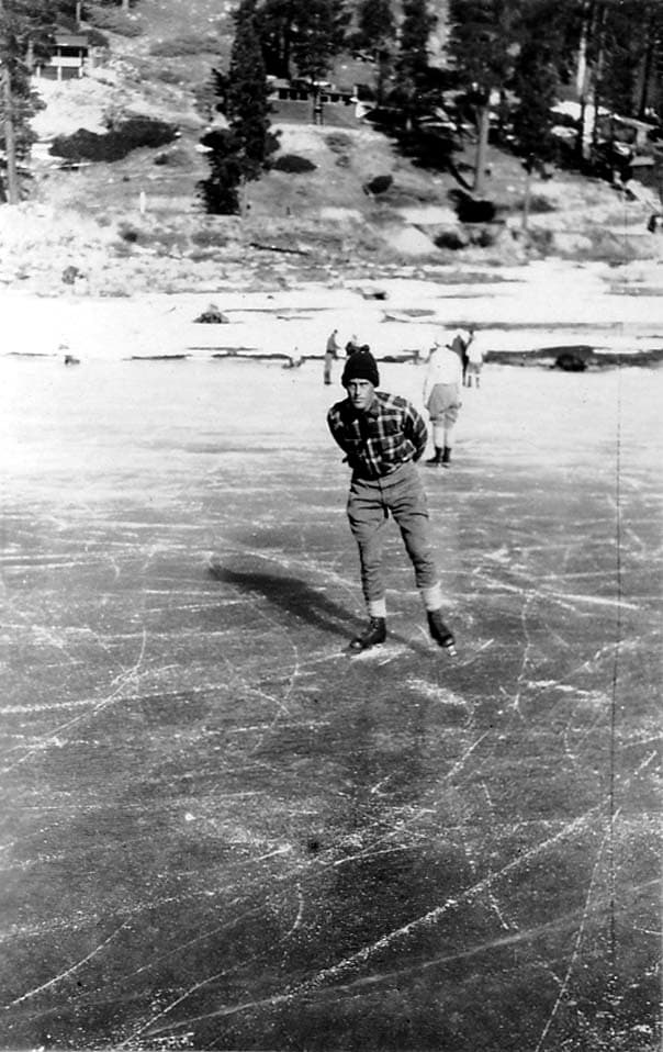 A self-portrait of Donald A. Hall Sr. wearing a plaid jacket and a knit cap, gliding toward the camera on a frozen lake with his hands clasped behind his back.