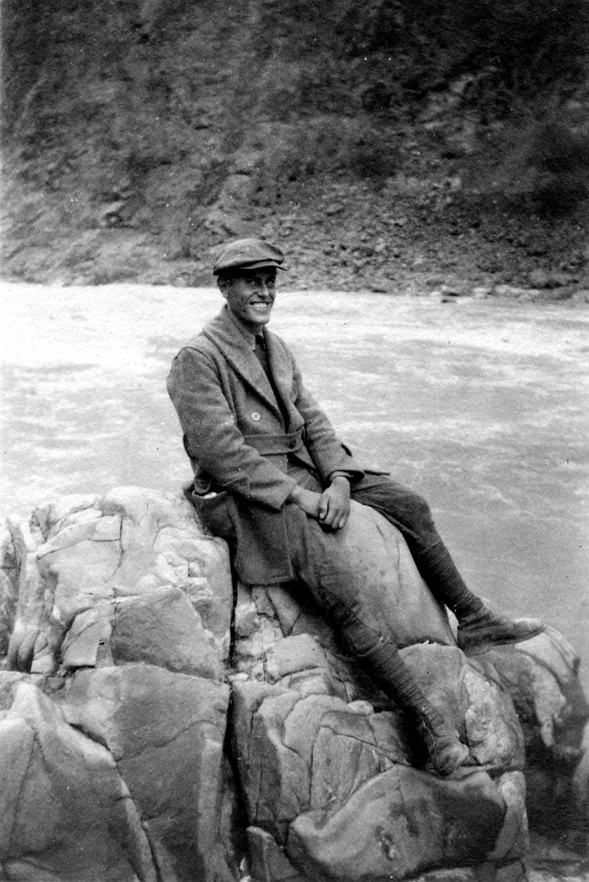 A smiling Donald A. Hall Sr. sitting on massive rocks beside a rushing river, wearing a heavy overcoat, tall boots, and a flat cap.