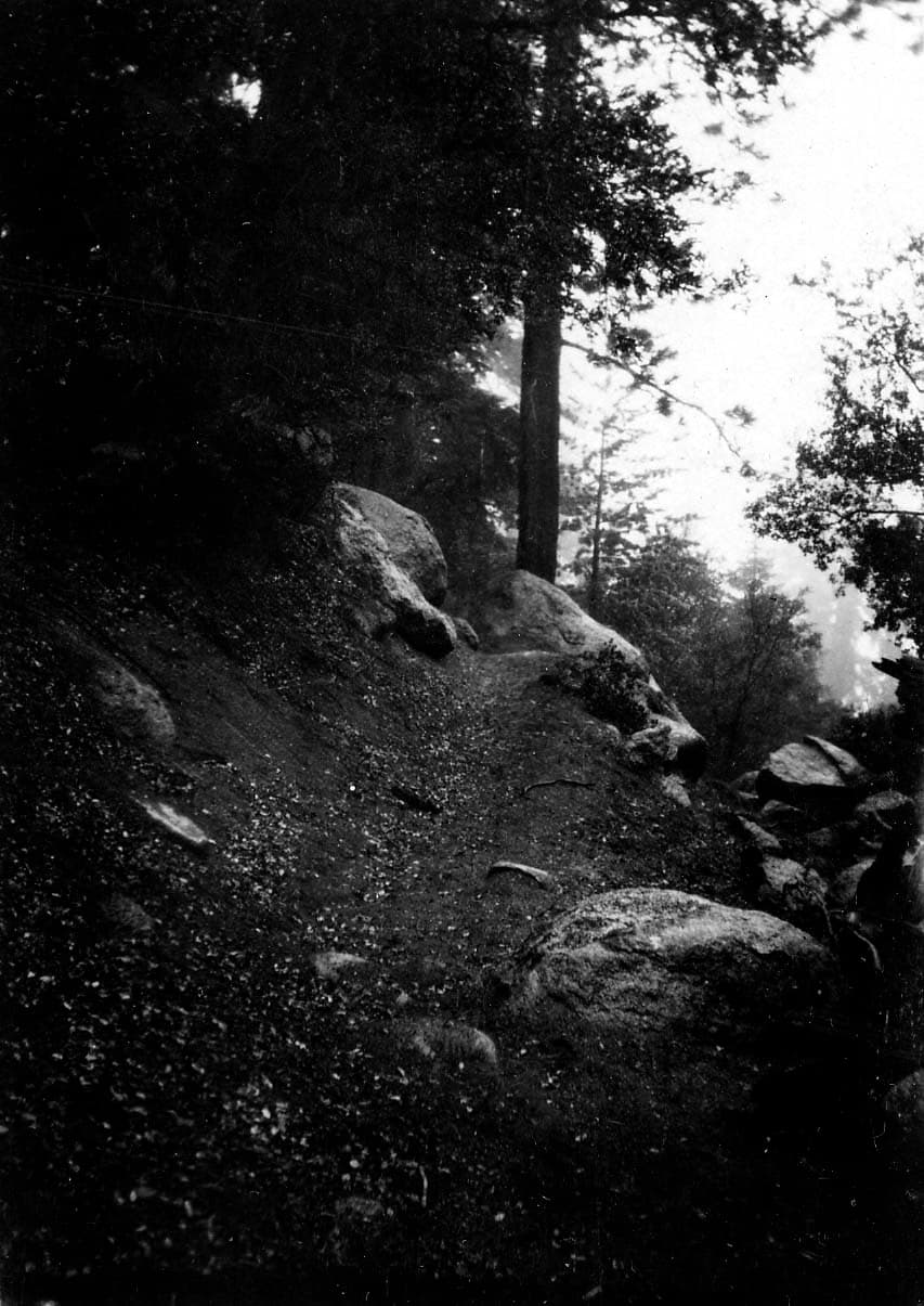 A steep, narrow dirt trail winding upward through dense shadow past large granite boulders and towering conifers.