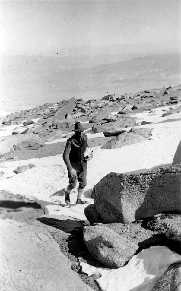A hiker wearing a fedora and carrying a bundle navigating a rocky, snow-covered mountain summit.