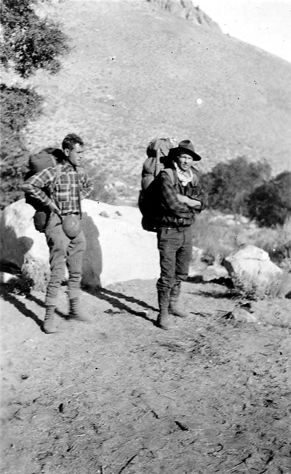 Two hikers wearing heavy canvas backpacks and rugged outdoor gear standing on a dirt trail in a mountain landscape.