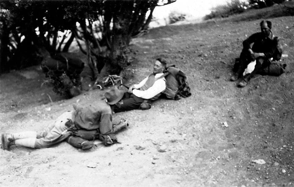Three hikers with massive canvas backpacks sitting and reclining in the dirt on a steep hillside to recover.