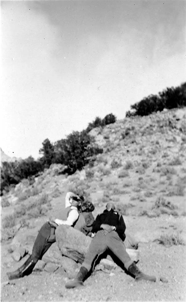 Three hikers, one leaning back against a large rock and two slumped forward in exhaustion, resting on a rocky mountain slope.