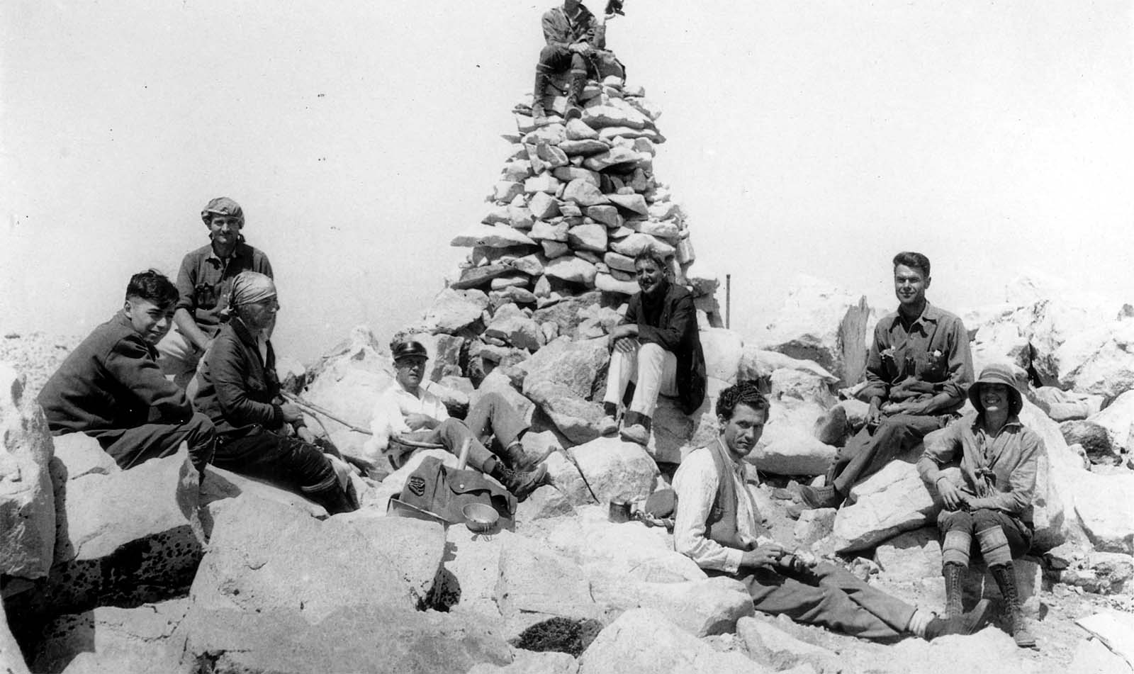 A group of eight hikers resting on rocks around a tall, man-made stone cairn on a mountain summit, with one hiker sitting at the very top.