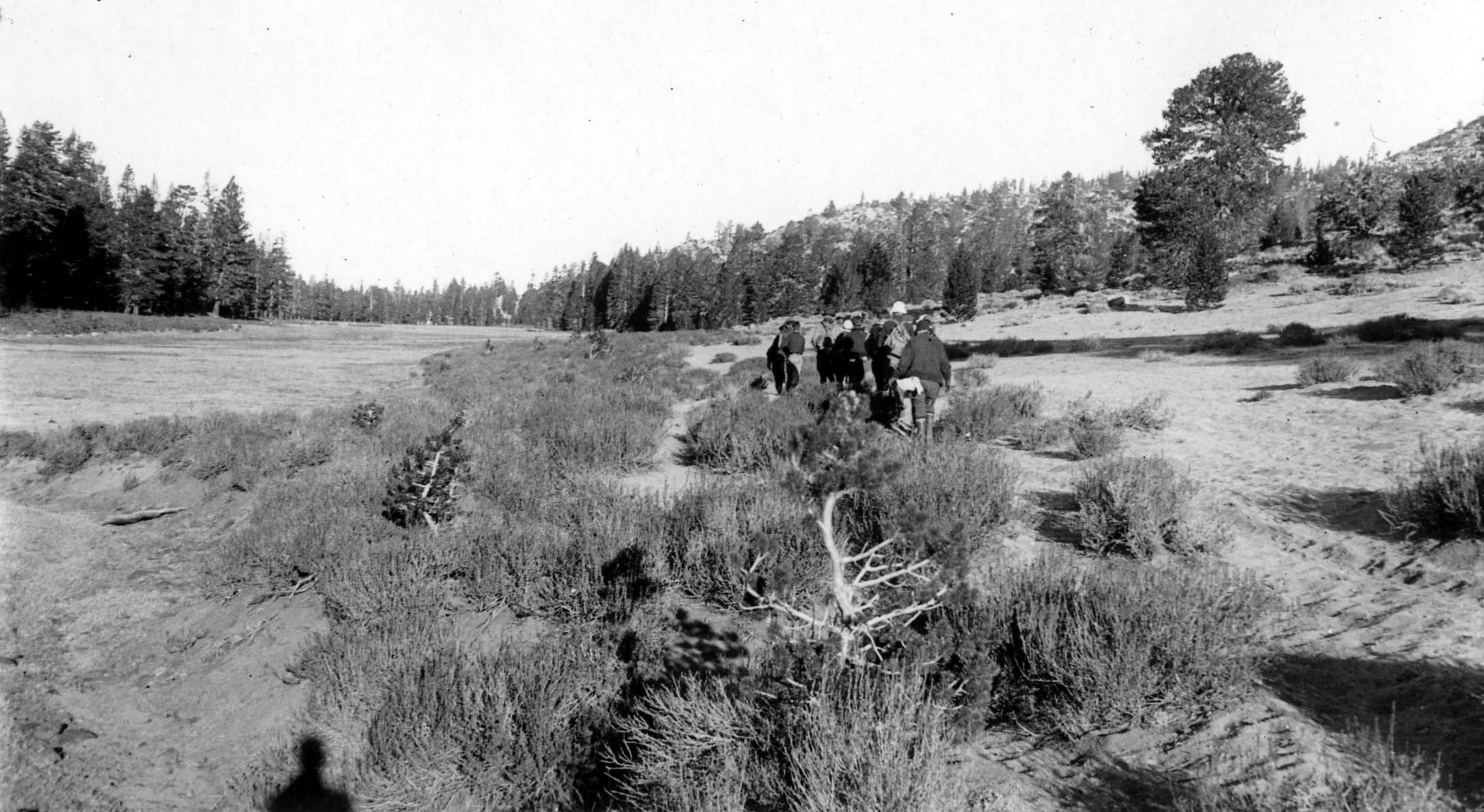 A line of hikers walking away from the camera down a sandy trail cutting through a wide, scrub-covered mountain valley.