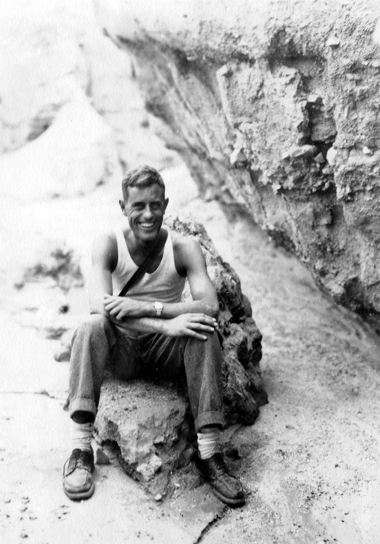 A smiling Donald A. Hall Sr. sitting on a rock beneath a rugged stone overhang, wearing a white tank top and hiking trousers.