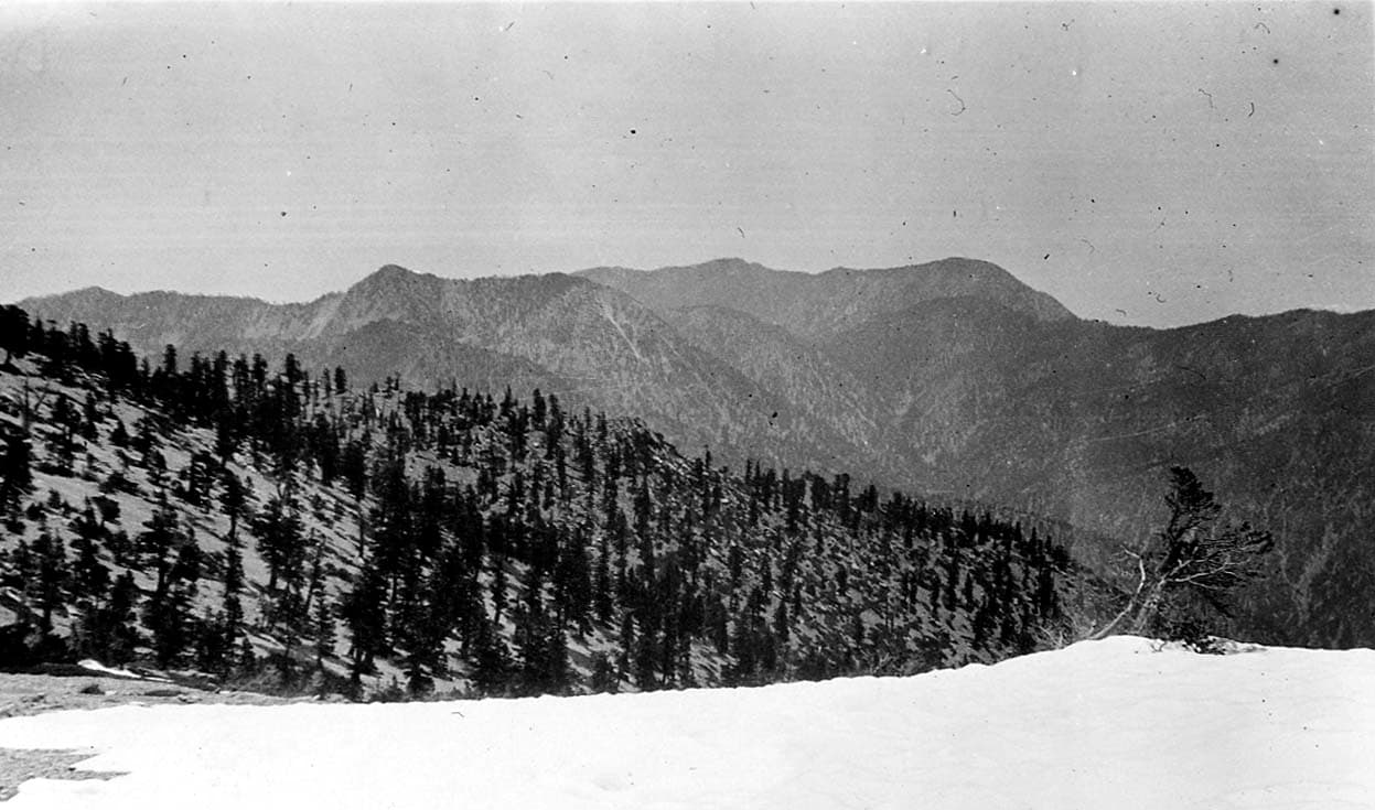 A wide landscape view of a rugged, snow-dusted mountain ridgeline stretching across the horizon under a cloudy sky.
