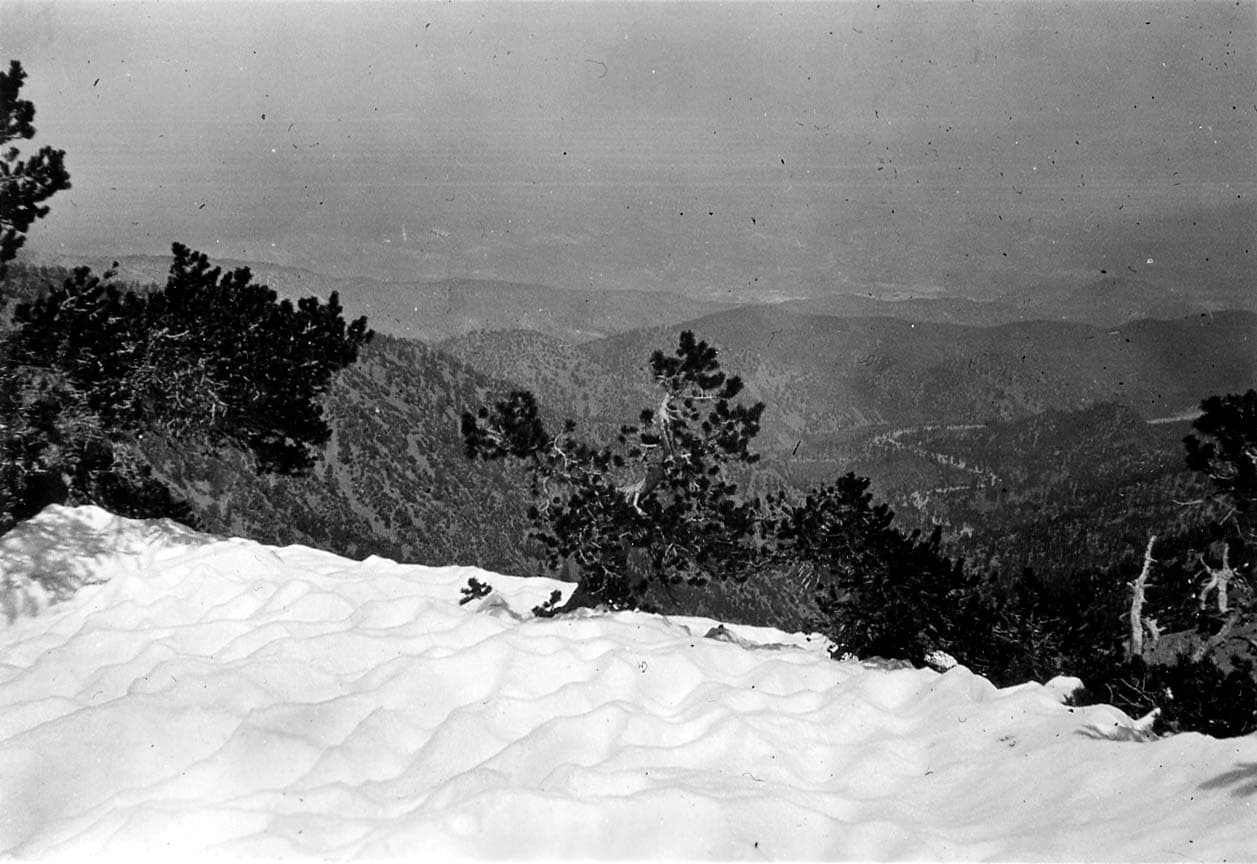 A high-altitude lookout over a vast, snow-dusted mountain valley, framed by dark pine branches in the foreground.