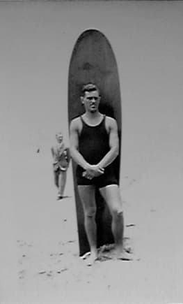 Donald A. Hall in tank swimsuit standing with a 10-foot solid wood surfboard on a San Diego beach.