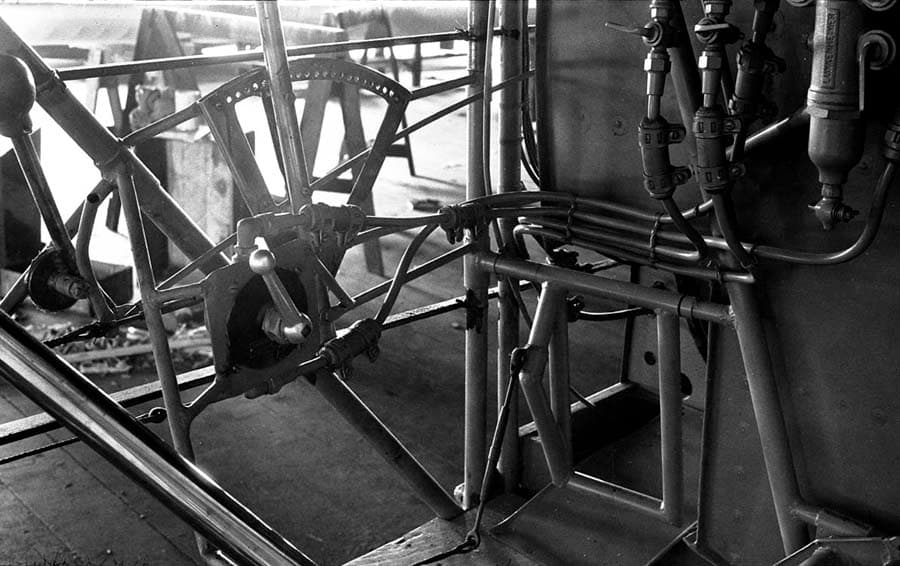 A close-up view inside the raw tubular steel cockpit of the Spirit of St. Louis, focusing on the mechanical flight controls and fuel routing.