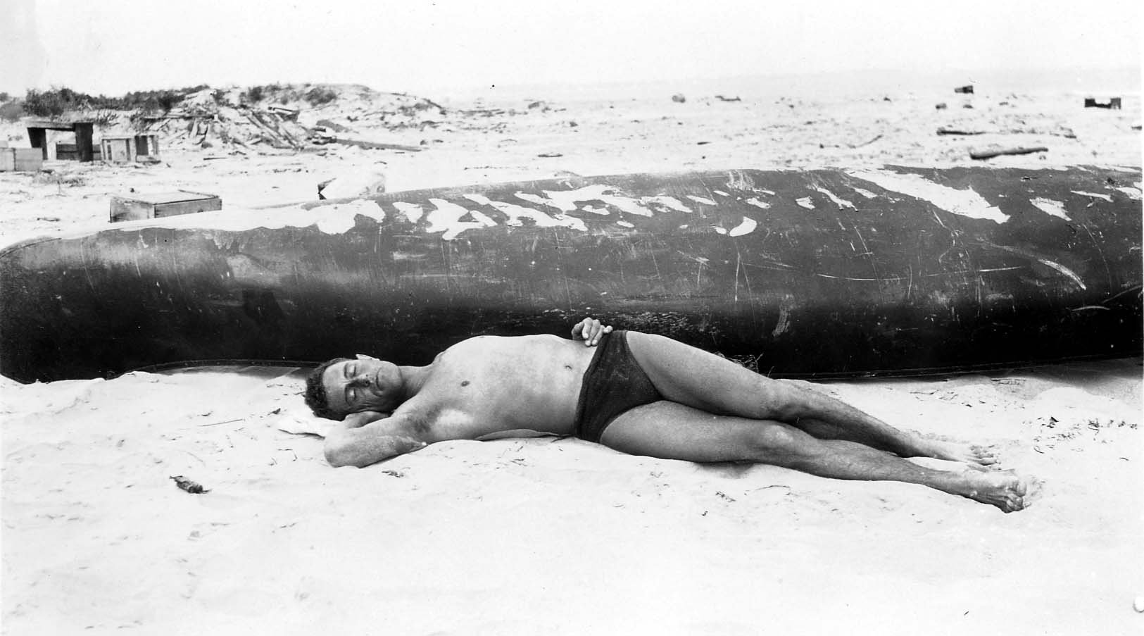Donald A. Hall Sr. sleeping or sunbathing on a sandy beach in dark swim trunks, lying next to the overturned hull of a dark canoe.
