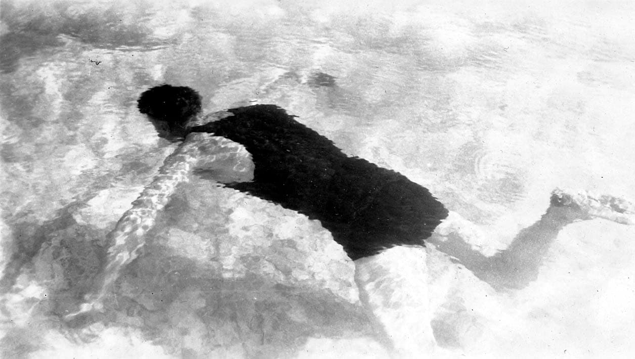 A high-angle photograph looking down through the surface of the water at a man gliding just beneath the surface.
