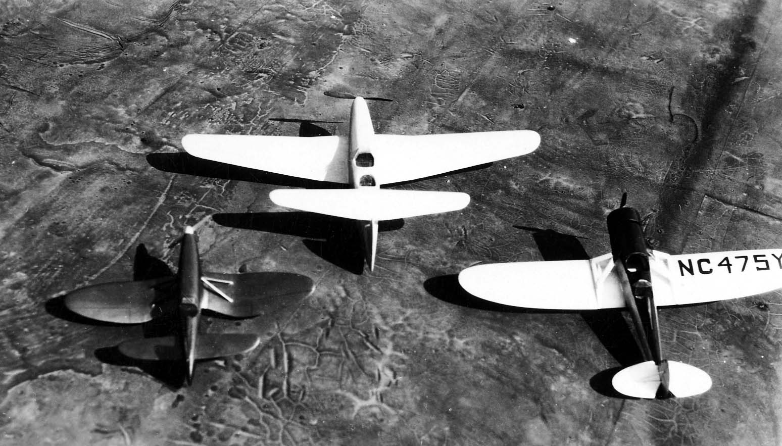 Three carved wooden aircraft models, including an experimental tandem wing design, resting on the textured roof of the aviation factory.