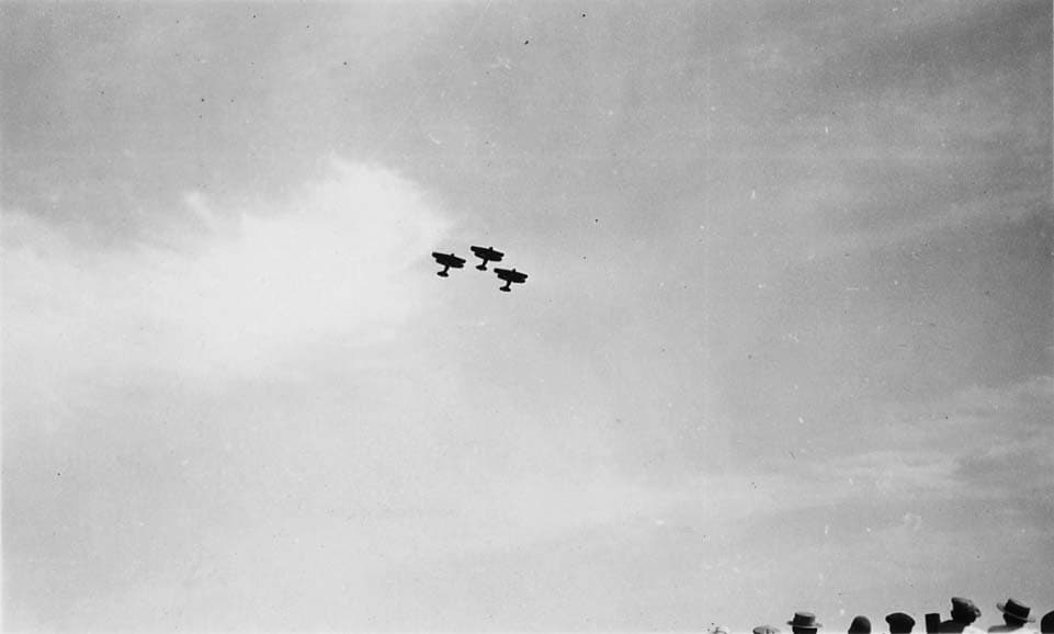 A very distant, high-altitude view of three biplanes flying in a tight V-formation against a bright sky.