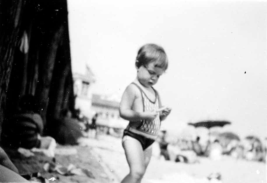 A young toddler in a vintage knit bathing suit standing on the sand examining a small discovery, with blurred beachgoers and umbrellas in the background.