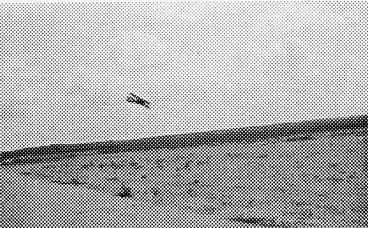 A grainy halftone print of a training biplane executing a low-altitude pass over the flat dirt airfield at Brooks Field.
