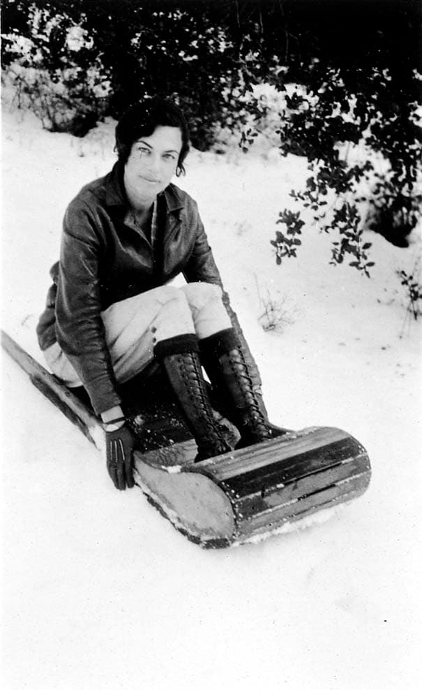 A portrait of an unidentified woman with a subtle smile, wearing a leather jacket, riding breeches, and tall boots while sitting on a wooden sled in the snow.