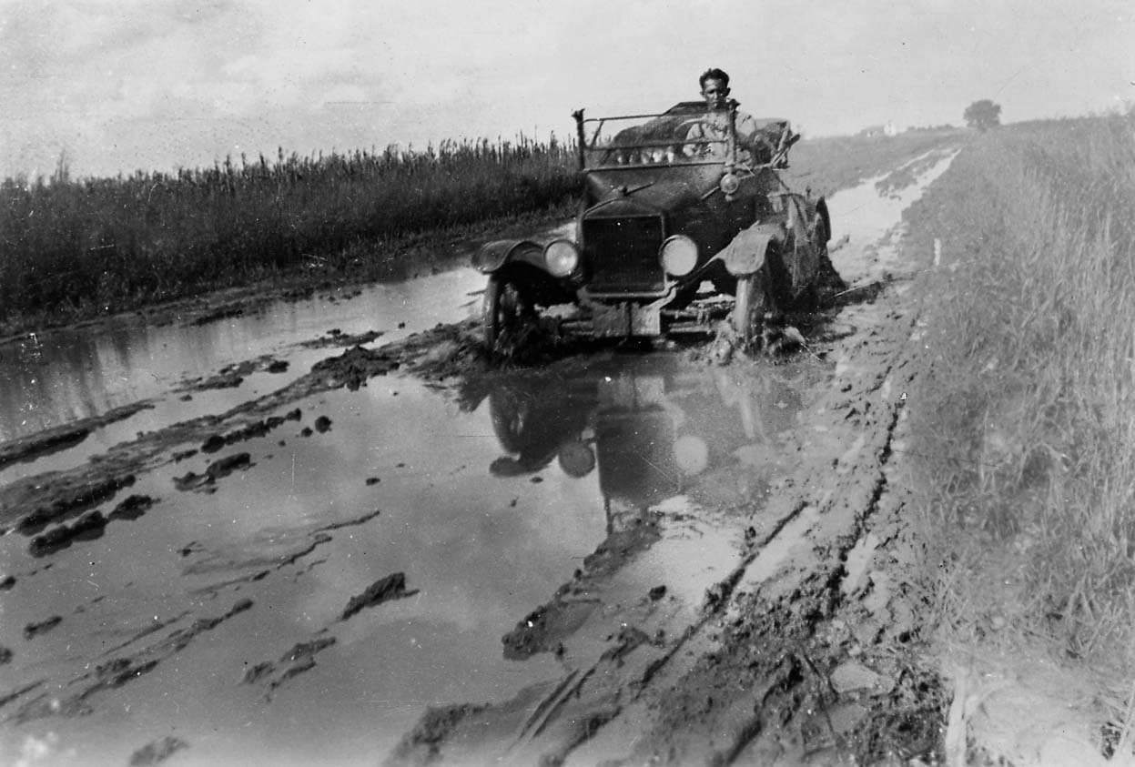 A vintage open-cab automobile, likely a Model T, buried to its axles in a deep, water-filled mudhole on a rural dirt road.