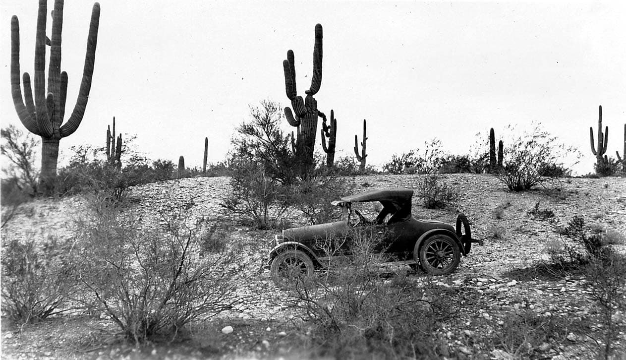 A vintage automobile parked off a dirt road in a rugged desert landscape, surrounded by scrub brush and towering saguaro cacti.