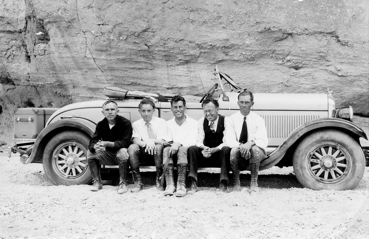 Five men sitting on a vintage convertible parked against a sheer rock wall in an alternate exposure of a timed photograph.