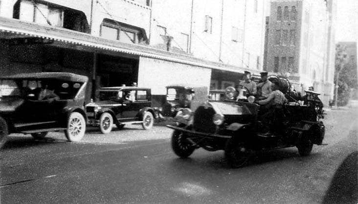 A vintage, open-cab fire engine being driven down a city street by uniformed firemen, passing parked 1920s automobiles.