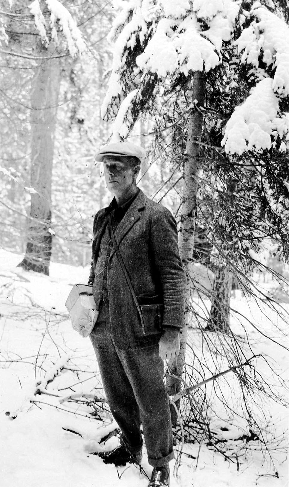 A man in a wool tweed three-piece suit, flat cap, and shoulder bag standing among heavy snow-laden conifer branches in a winter forest.