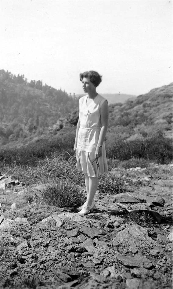 A full-length outdoor portrait of a young woman in a sleeveless drop-waist 1920s summer dress standing on a rocky mountain ridge dotted with chaparral.