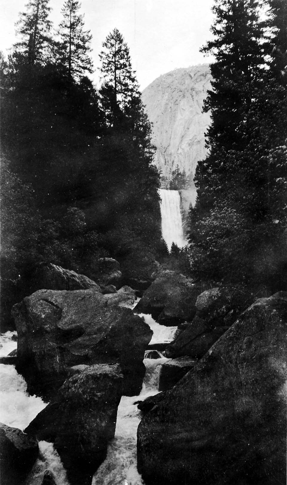 A powerful rectangular waterfall cascading into a deep granite canyon, viewed down a boulder-strewn rushing river framed by tall conifers.