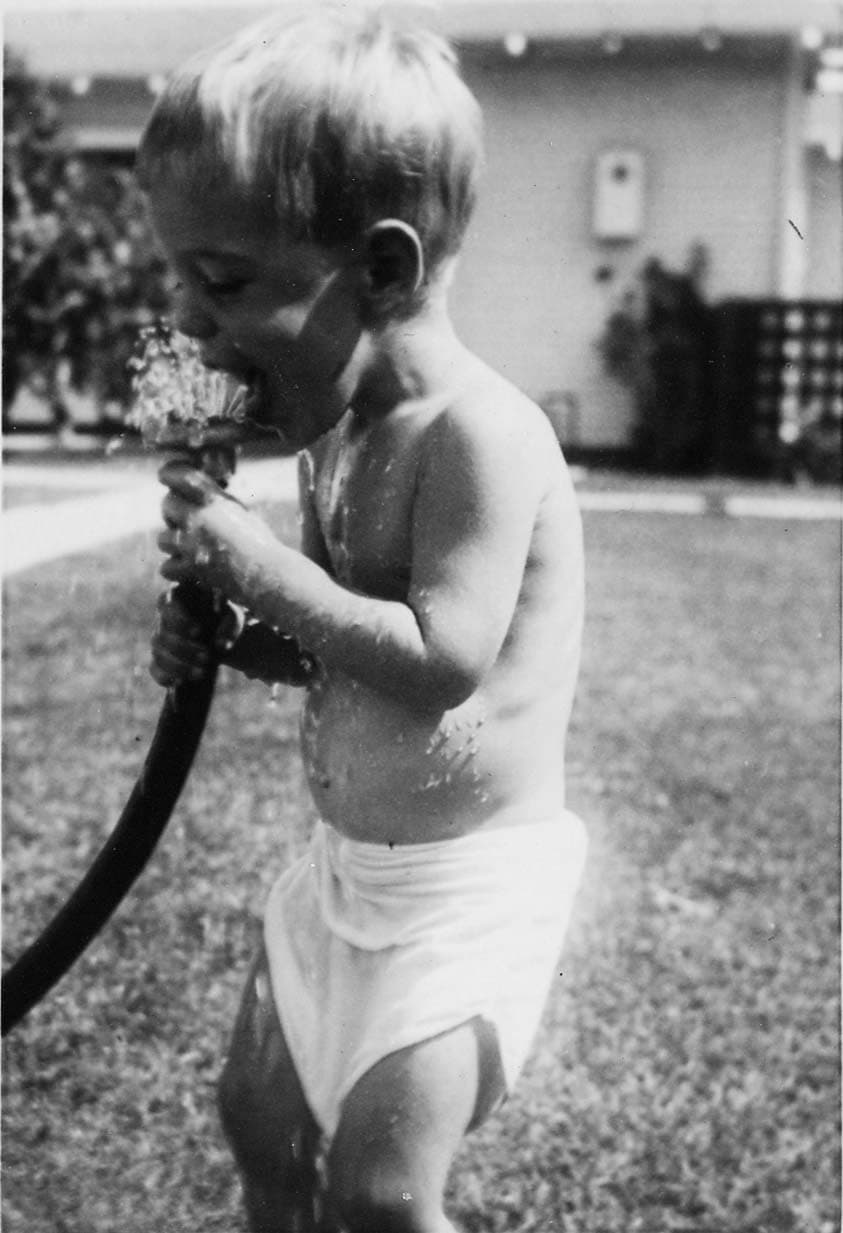 A candid photograph of a young toddler boy in a diaper playfully drinking water directly from a running garden hose in a grassy yard.