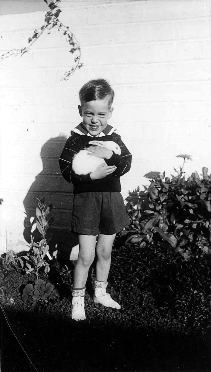 A young boy, Don Hall Jr., standing in front of a white brick wall and bushes, smiling while gently holding a fluffy white rabbit.