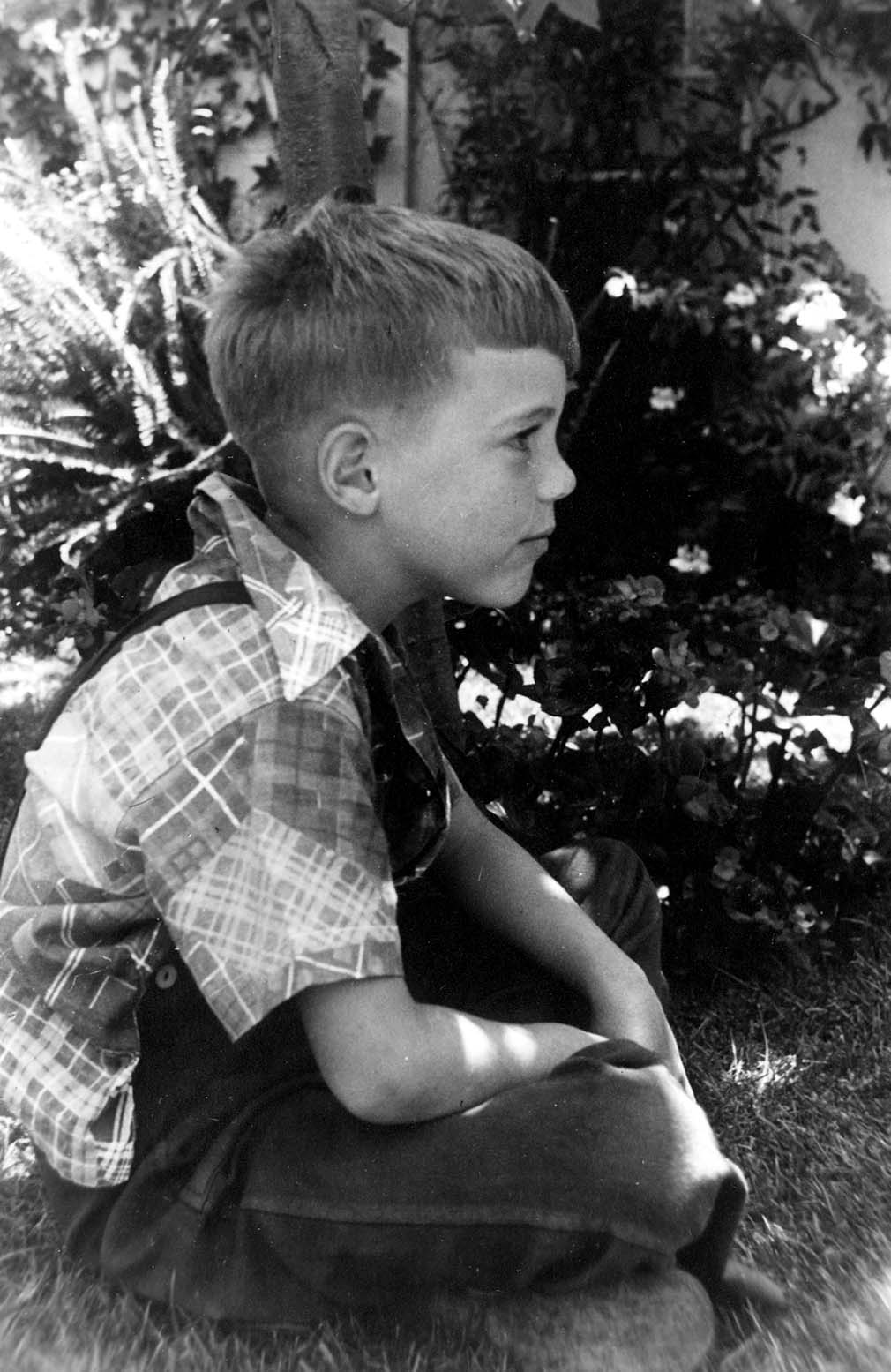 A young boy, Don Hall Jr., sitting quietly in the grass in a plaid shirt and overalls, looking off to the right in profile.