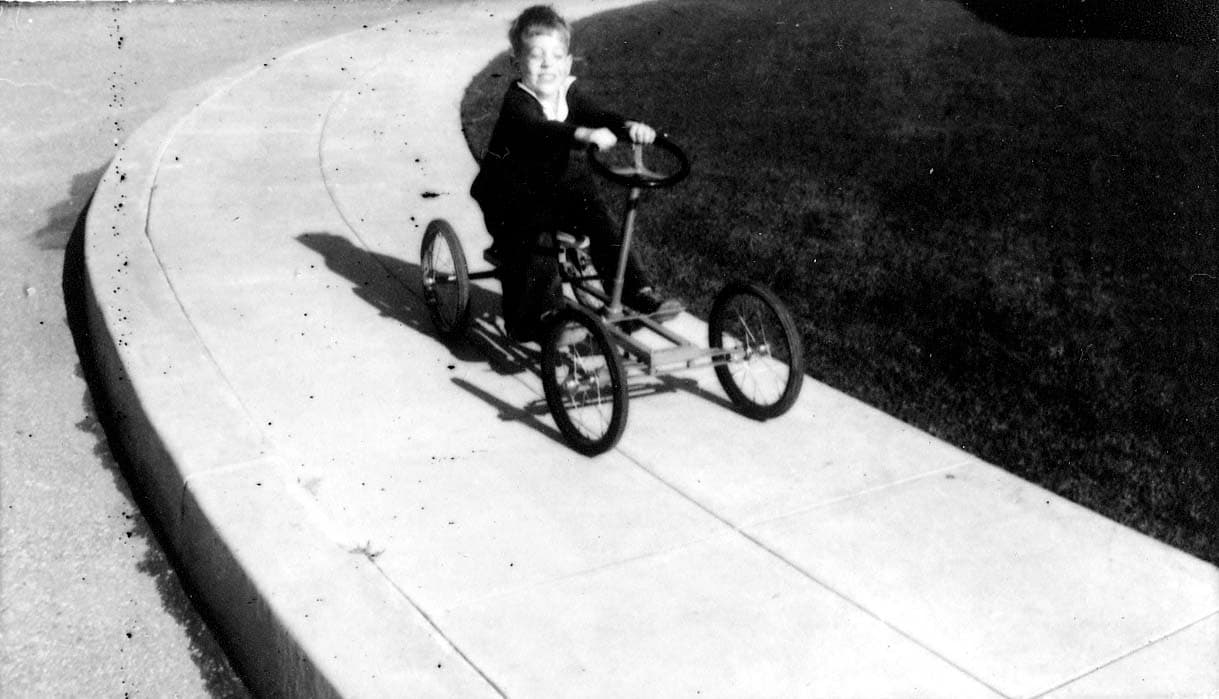 A young boy, Don Hall Jr., smiling broadly as he steers a heavy, four-wheeled pedal car down a curved concrete sidewalk.