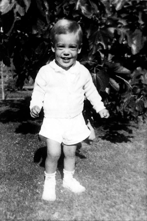 A toddler, Don Hall Jr., standing on a lawn wearing a white sweater and shorts, smiling brightly at the camera.