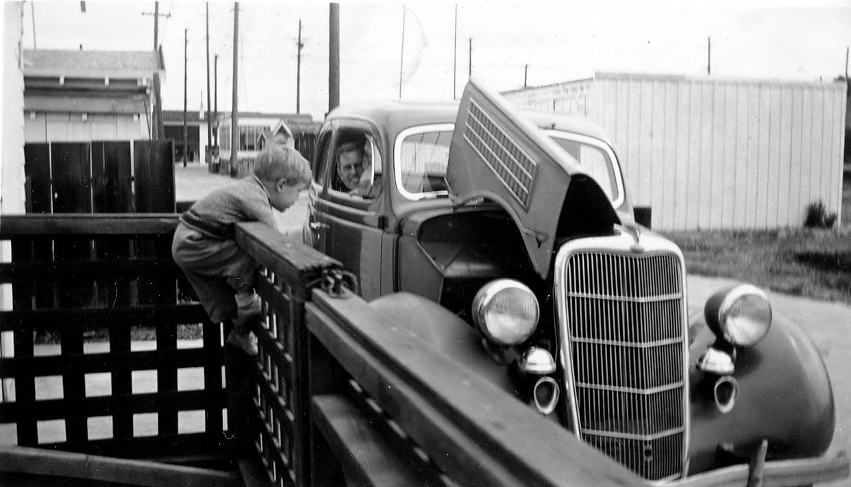 Donald Hall Sr. looking from the window of a 1935 Ford coupe with open hood while young Don Jr. climbs a porch railing.