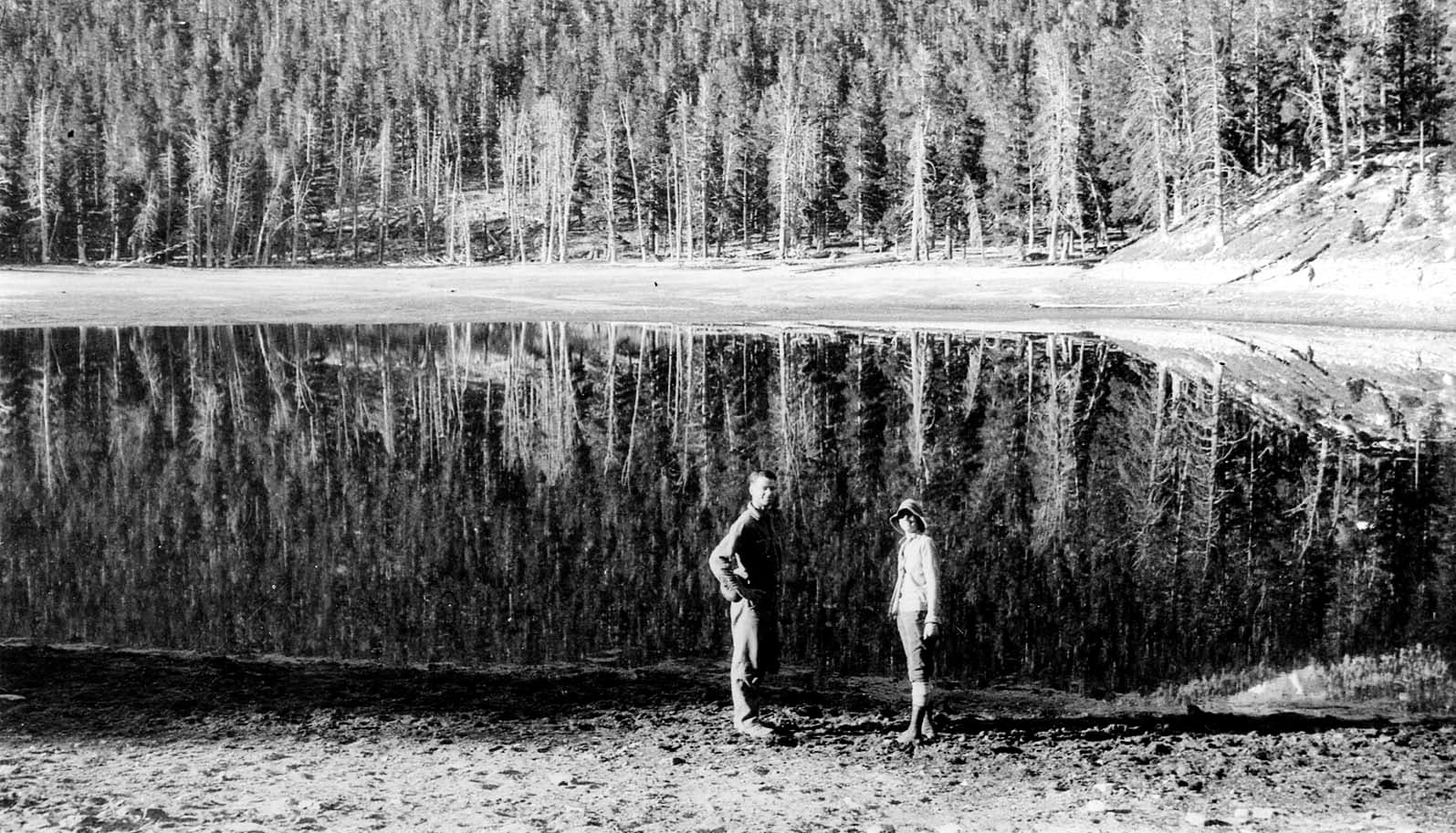 Two hikers standing on the sandy shore of Lake Cuyamaca with pristine, mirror-like reflections of tall pines across the water.