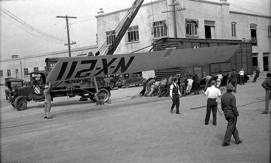 The 46-foot wing of the Spirit of St. Louis suspended by a mobile crane truck outside the Ryan factory with ground crew holding tether lines.