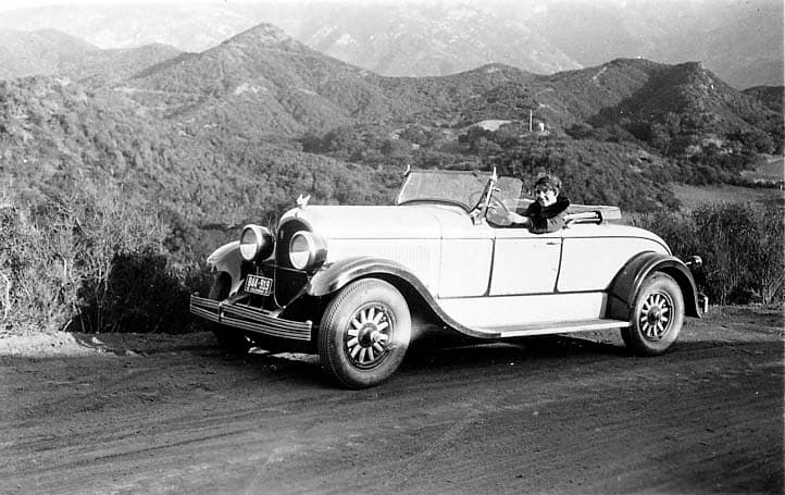 Donald A. Hall Sr.'s 1925 Studebaker Roadster parked on an unpaved mountain road with a smiling woman behind the wheel against layered hills.