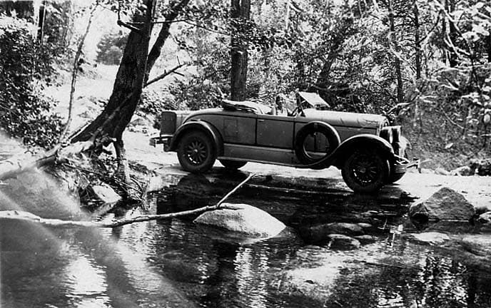The 1925 Studebaker Roadster parked on a low-water concrete bridge over a rocky, flowing woodland creek beneath shaded trees.