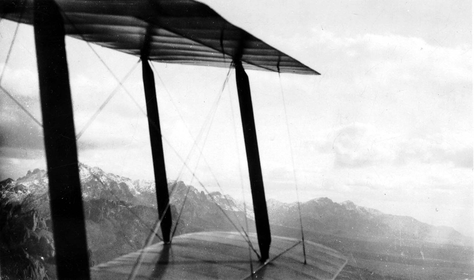 An aerial view looking out past the struts and fabric wings of a biplane, with snow-dusted mountains in the distance.