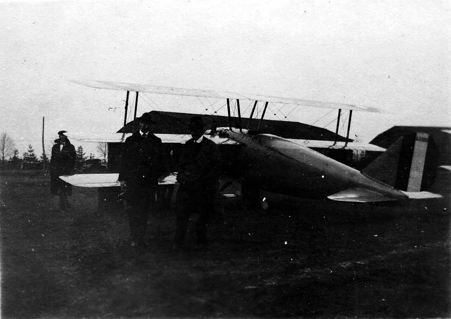 Two men dressed in 1920s overcoats and fedoras standing in front of a sleek biplane with an enclosed fuselage and striped military rudder.