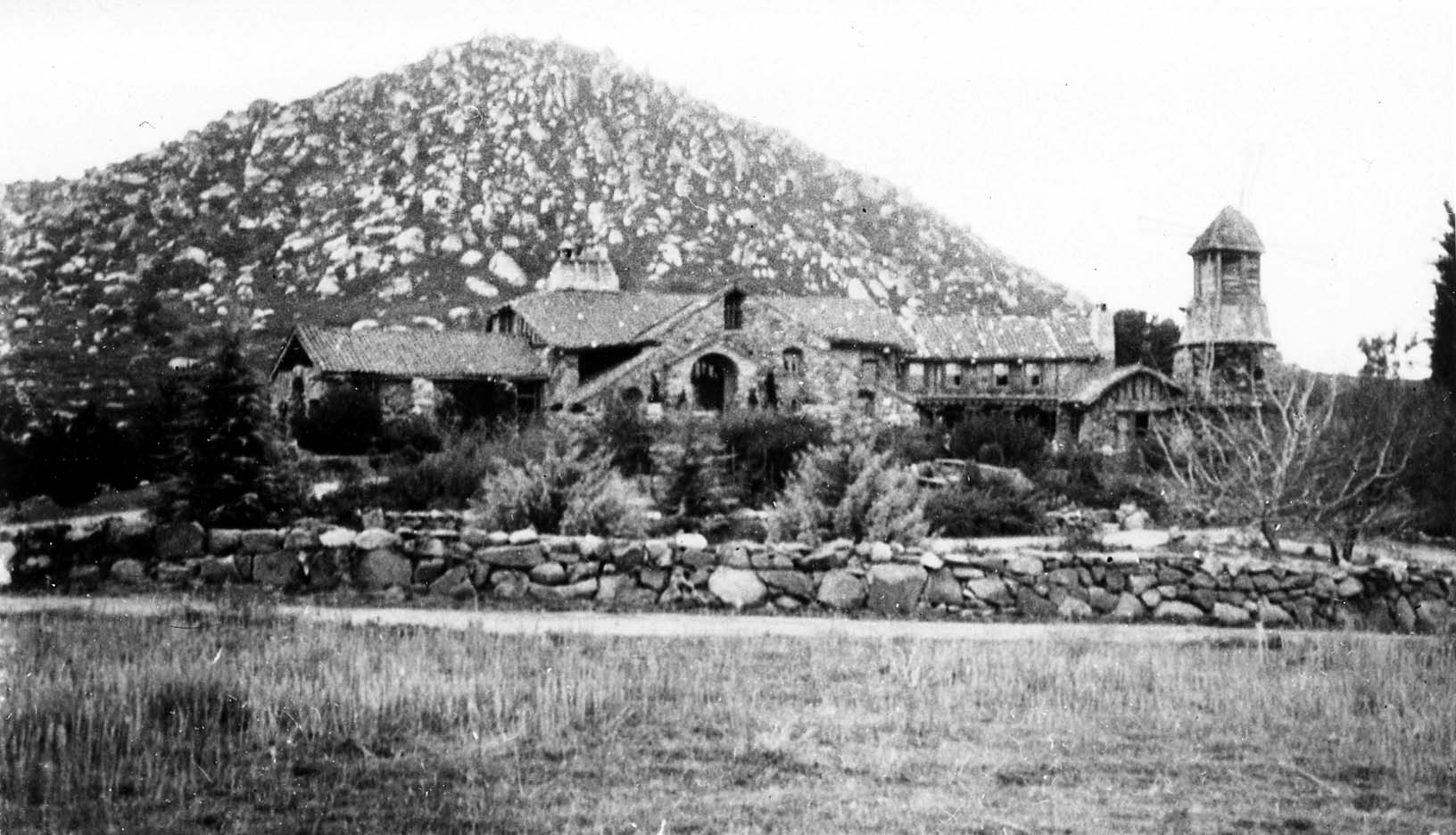 A sprawling granite stone castle with Spanish tile roofs, arched entryways, and a wooden windmill tower at the foot of boulder-strewn Mount Woodson.