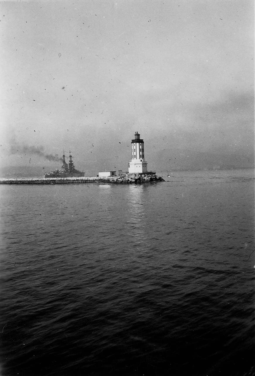 A view across Los Angeles Harbor toward the historic Angels Gate Lighthouse at the end of the breakwater, with a U.S. Navy cage-mast battleship steaming in the distance.