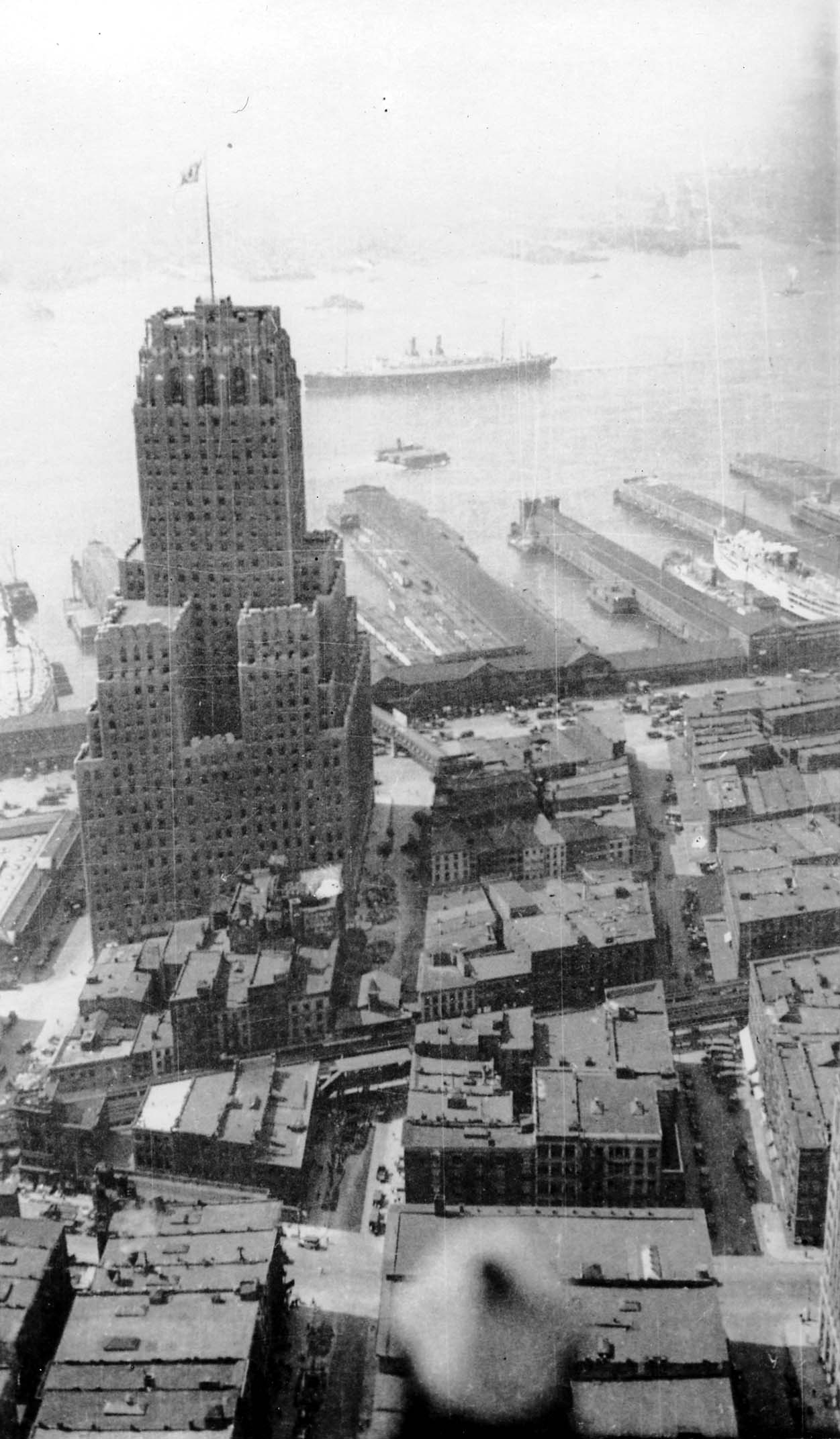 A high-angle view looking west from the Woolworth Building over the Art Deco Barclay-Vesey Building toward Hudson River piers and an underway ocean liner.