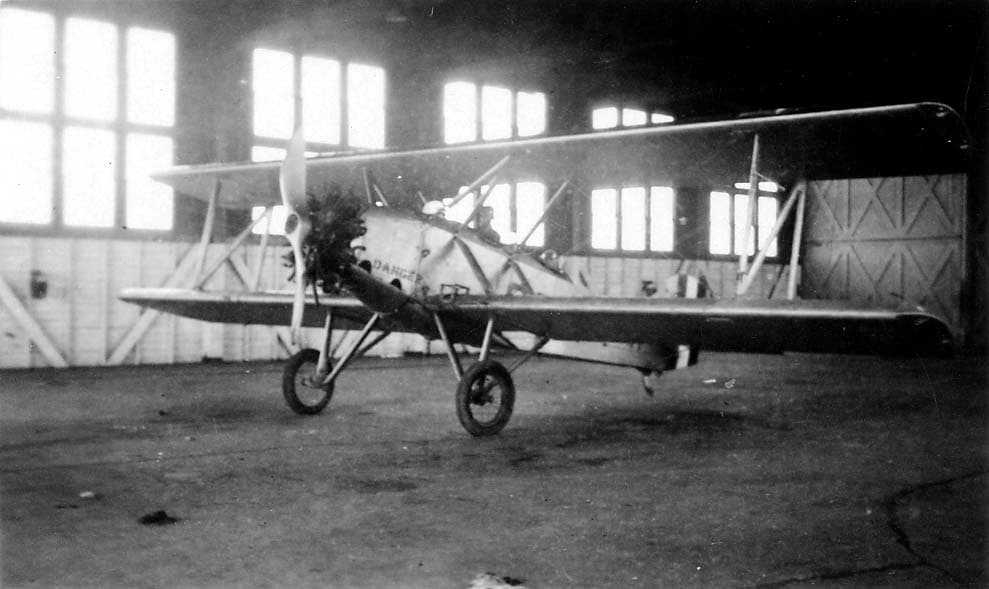 An open-cockpit biplane inside a brightly lit hangar with the word DANGER stenciled in bold block letters near the propeller arc.