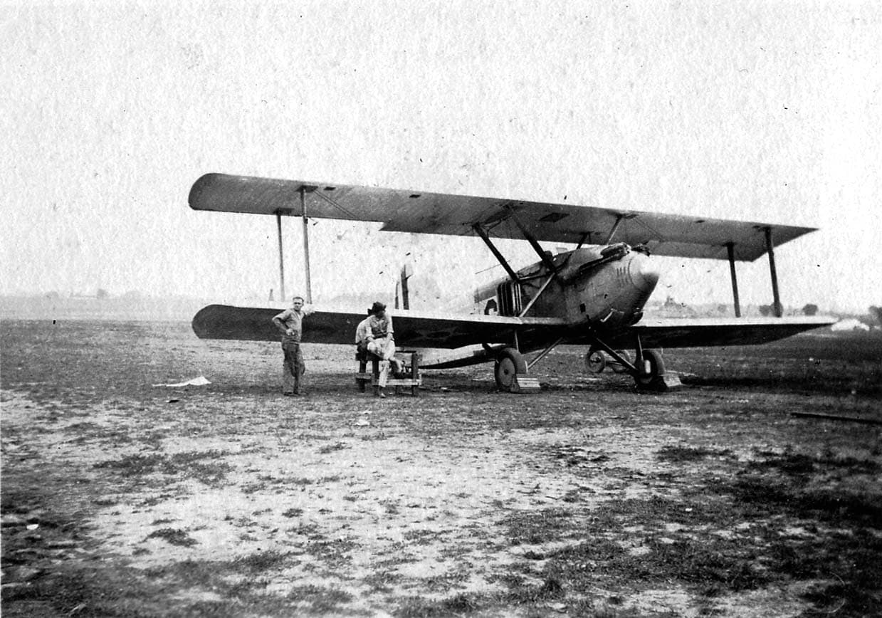 Ground crew mechanics resting on a tool chest beneath the lower wing of an experimental pursuit biplane with a streamlined nose spinner and tunnel radiator.