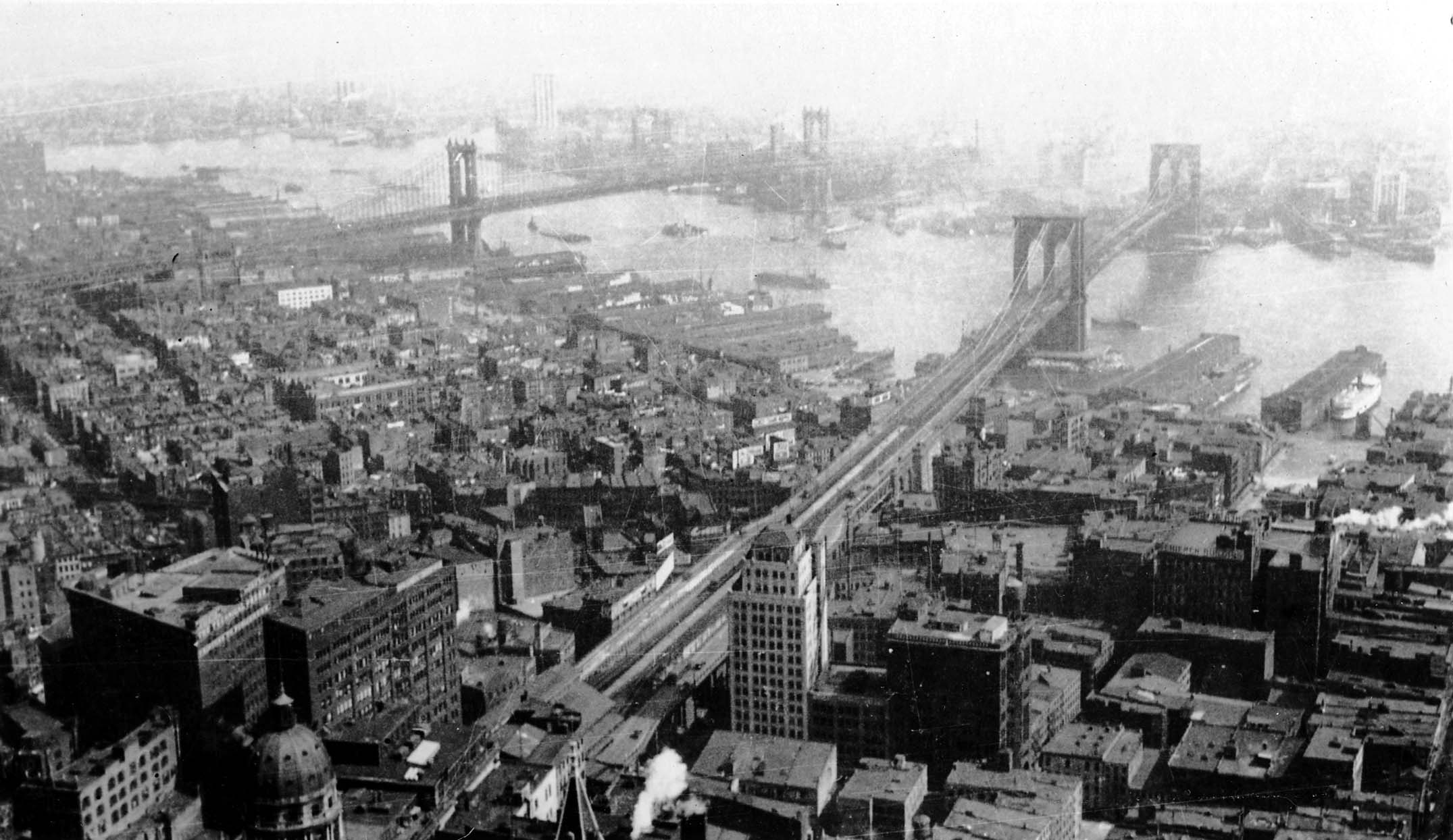 A panoramic vista from the Woolworth Building showing the Brooklyn and Manhattan bridges spanning the East River, with the copper dome of the World Building in the foreground.