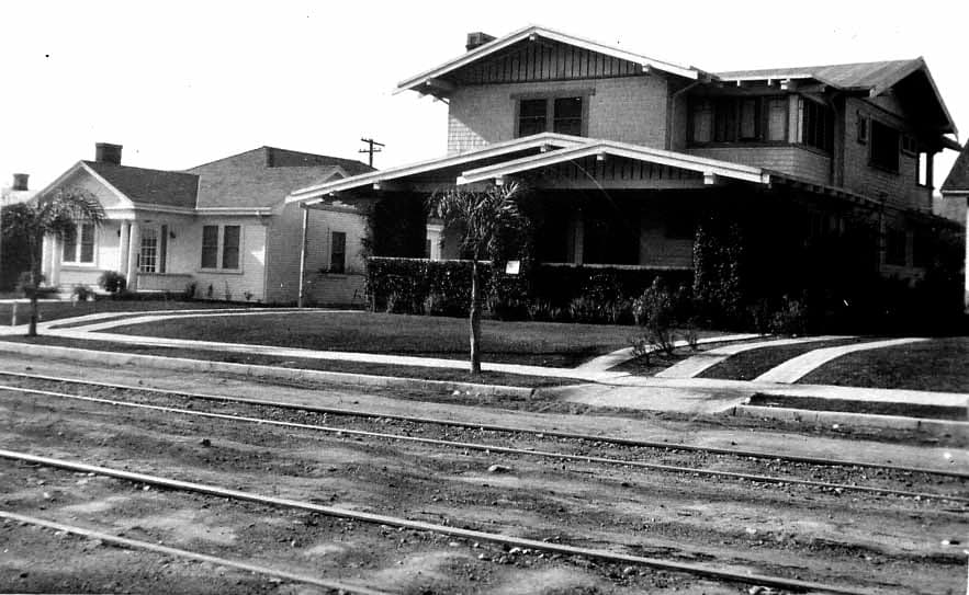 A two-story California Craftsman airplane bungalow with deep gabled eaves, wide front porch, and unpaved streetcar tracks in the foreground.