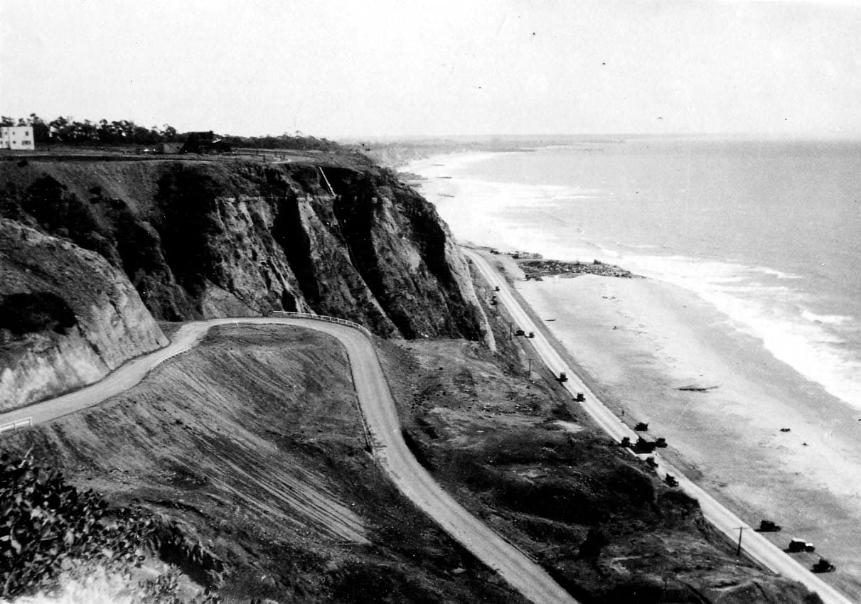 A sweeping elevated view of the curving California Incline descending the Santa Monica Palisades to the coast highway, with vintage cars parked along the beach.