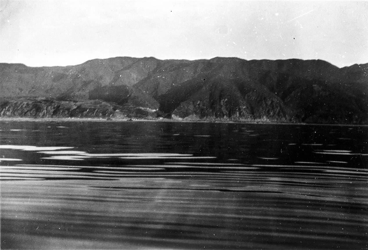 Calm ocean waters showing smooth glassy slick bands and gentle surface swells reflecting beneath steep coastal mountain slopes.