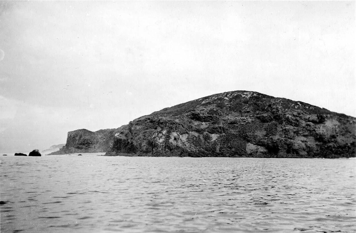 A rugged, volcanic coastal headland with steep rocky cliffs and small offshore sea stacks rising from a gentle ocean swell.